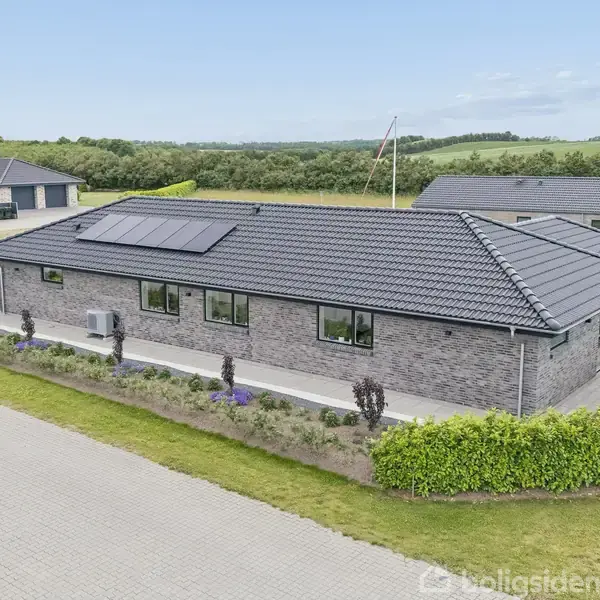 Et moderne, en-etagers hus med solpaneler på taget står omgivet af en grøn have. En asfalteret sti løber langs huset i et fredeligt villakvarter med grønne marker i baggrunden.
