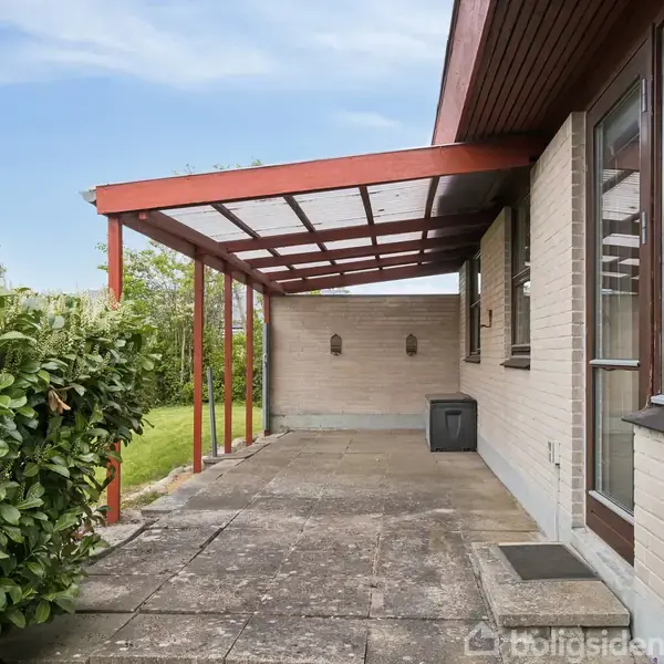 En overdækket terrasse lavet af lyse mursten og røde træbjælker i en have, omgivet af hæk og græsplæne. Siden af huset har store vinduer.