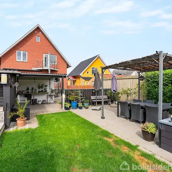 Et hus i røde mursten står i baggrunden med to andre huse. På terrassen foran er der sorte havemøbler under en pergola ved siden af en lille have med græs og planter.