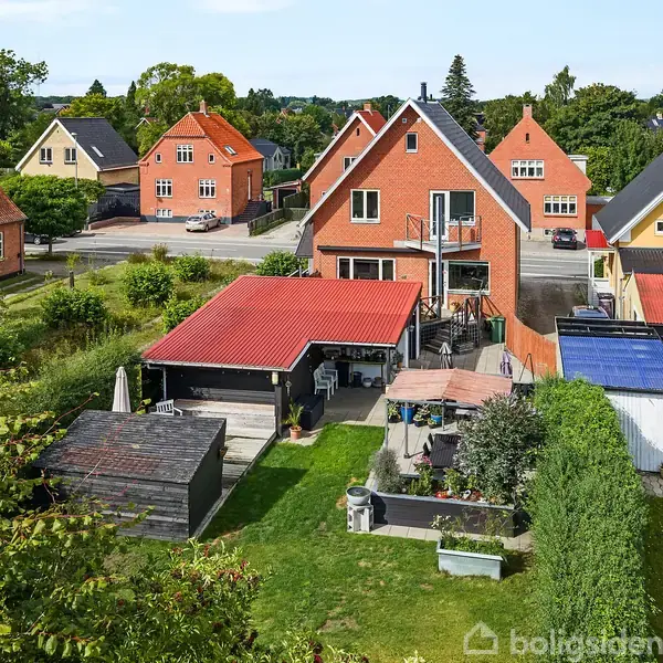 En rød murstenshus med skråt tag står i et villakvarter. Omkring huset ses en have med grønne træer og buske, og der er en carport med et rødt tag foran huset.