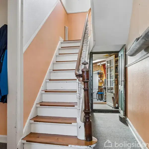 Trætrappe fører opad ved siden af beige væg. En dør ved trappens fod åbner ind til et værksted med værktøj og hylder.