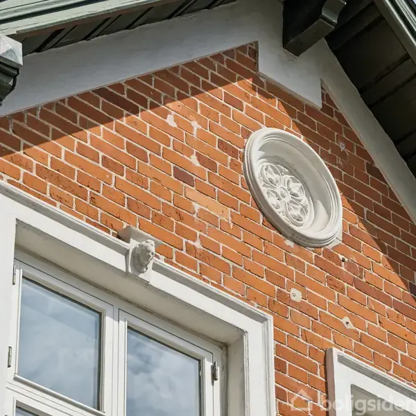 Murstensfacade med et cirkulært, dekorativt hvidt relief. Reliefet er over et vindue med hvide rammer. Sollys skaber skygger på murværket.