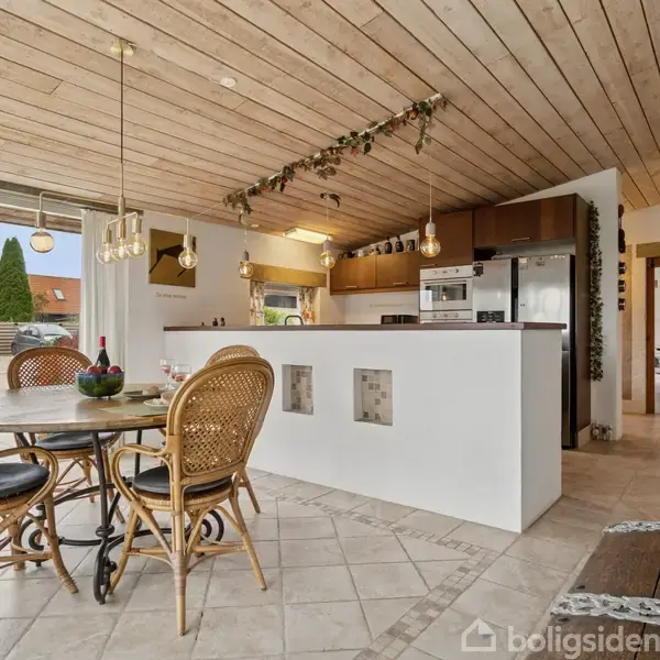 Rundt bord med fire kurvestole står på et flisegulv i et køkken. Køkkenet har hvide vægge, træloft og en halvmur med indbygget ovn og køleskab i baggrunden.