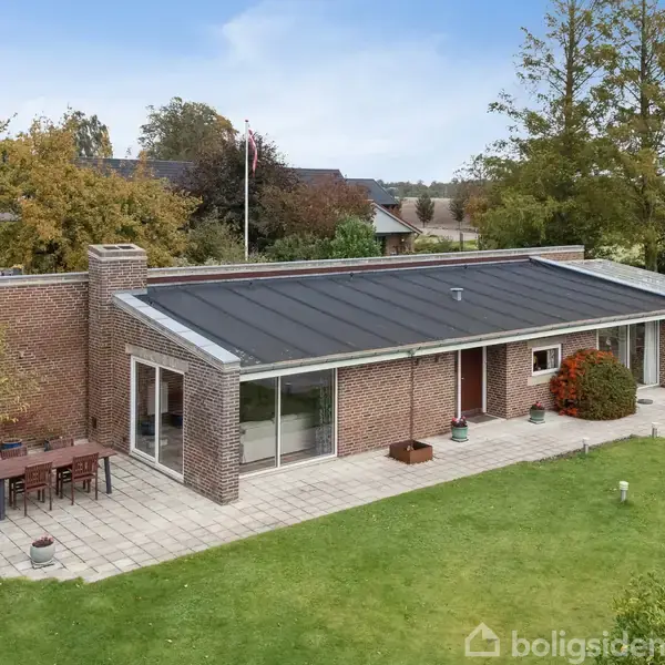 Et enkelt etplans murstenshus med fladt tag har en stor, flisebelagt terrasse. Det er omgivet af en velplejet græsplæne og grønne træer i en landlig have.