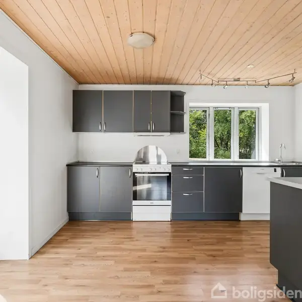 Køkken med grå skabe og indbygget ovn. Træloft og trægulv skaber en varm atmosfære. Store vinduer og en terrasse skaber lys og åbenhed. Intet tekst på billede.