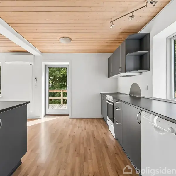 Køkken med grå skabe og træloft, har store vinduer og trægulv. En opvaskemaskine og ovn er integreret. Dør fører til en terrasse med udsigt til frodig have.