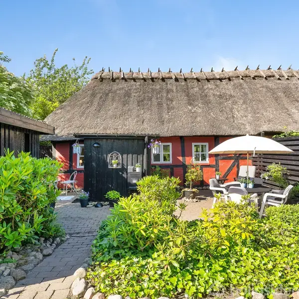 Stråtækt hus står i en hyggelig have med borde og stole under en parasol. Sollys skinner ned, og omgivelserne er frodige med grønne buske og stier af sten.