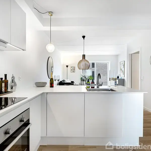 Et moderne køkken med hvide skabe og en indbygget ovn, dekoreret med flasker og skåle. En rund lampe hænger over den åbne køkkenø, omgivet af lyse trægulve.