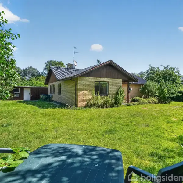 Et gult murstenshus med skråt tag står i en grøn have. Foran huset er der en græsplæne. Et bord og stole er placeret nær buske.