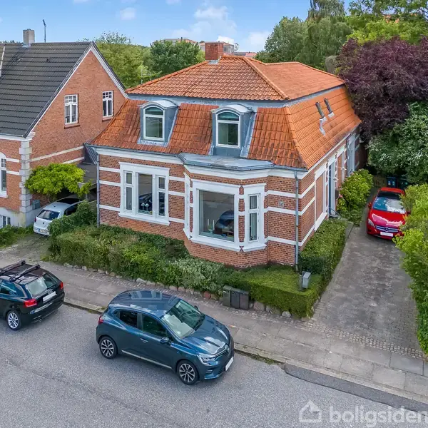 Et rødt murstenshus med rødt tegltag har to biler parkeret foran. Huset ligger i en række af lignende bygninger på en stille vej omgivet af buske og træer.