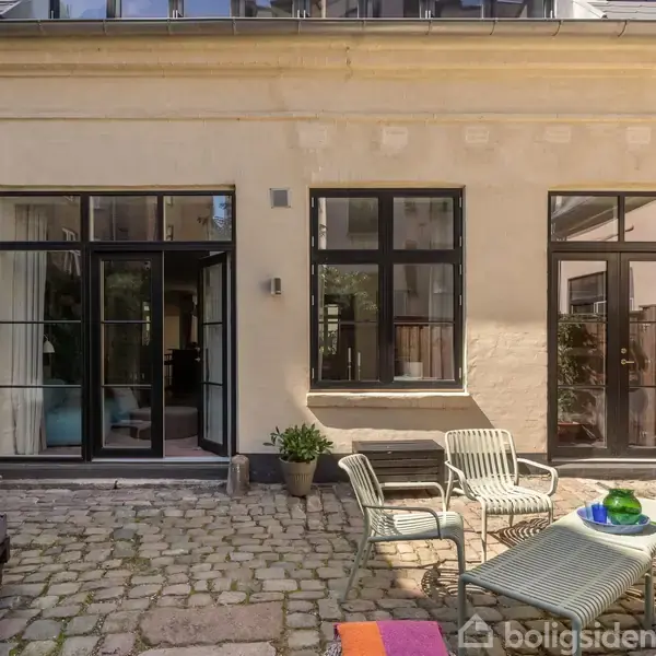 En brostensbelagt gårdhave med havestole og bord foran en beige bygning med store vinduer og glasdøre. En grøn plante står ved døren, og en omkringliggende mur omkranser området.