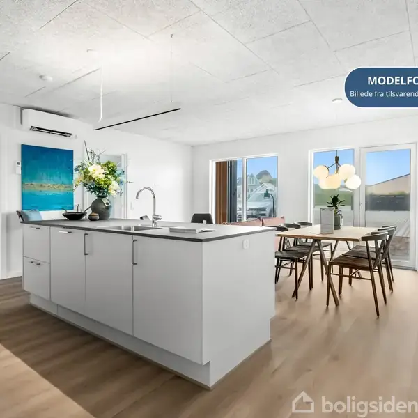 Køkken-ø med vask og blomster-arrangement står i moderne stue. Spisebord med seks stole står tæt på vinduer. På væggen hænger et kunstværk. Tekst: "MODELFOTO Billede fra tilsvarende lejlighed".