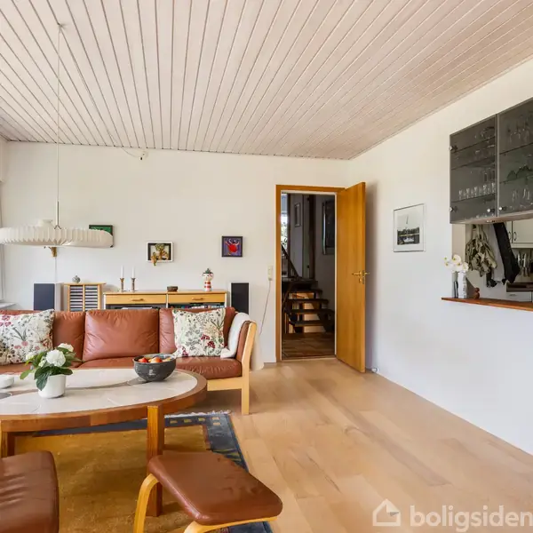 Stue med lædersofa og sofabord med blomster, ved siden af store vinduer. Træspisebord i baggrunden, åbning til køkken til højre, malerier på væggene og åbent dør til entreen.