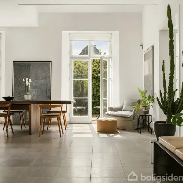 En stue med en åben pejs og ildsted. Et stort vinduesparti tillader lys at strømme ind, med en spisestue og en sofa til højre omgivet af grønne planter.