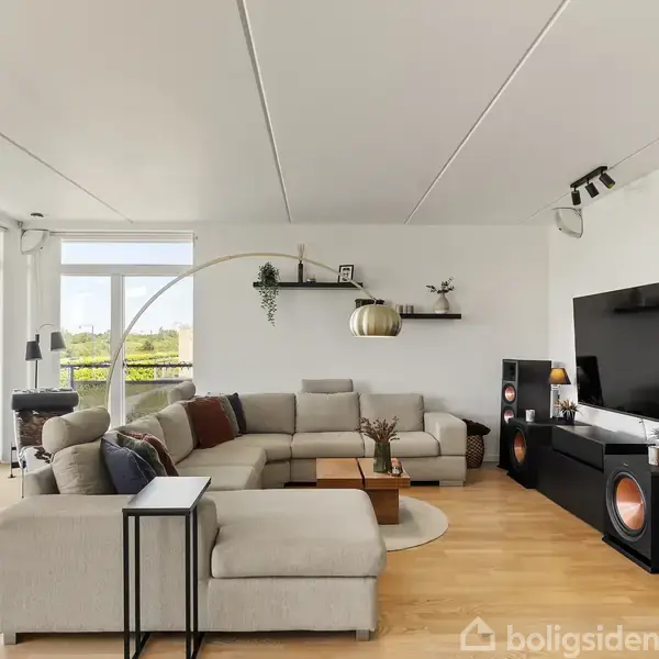 En stor L-formet sofa står i en moderne stue sammen med et sofabord. Et stort fladskærms-tv er placeret på en skænk, omgivet af højttalere. Stuen har store vinduer med udsigt.