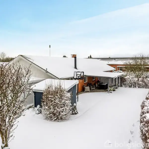 Et hus dækket af sne står stille i en vinterlig have omkranset af hegn og buske. Sneen dækker både taget og græsset, mens himlen er let skyfri.
