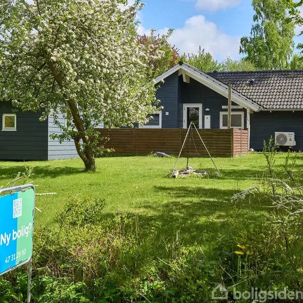 Et gråt træhus står i en frodig have med grøn græsplæne og blomstrende træer. Et skilt viser teksten "Ny bolig? 47 33 16 16."