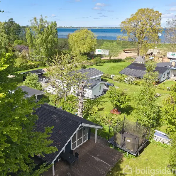 Sommerhus med sort tag ligger i frodig have med trampolin, omgivet af træer og buske. I baggrunden ses havet og flere bygninger. Scenen er belyst af solskin.