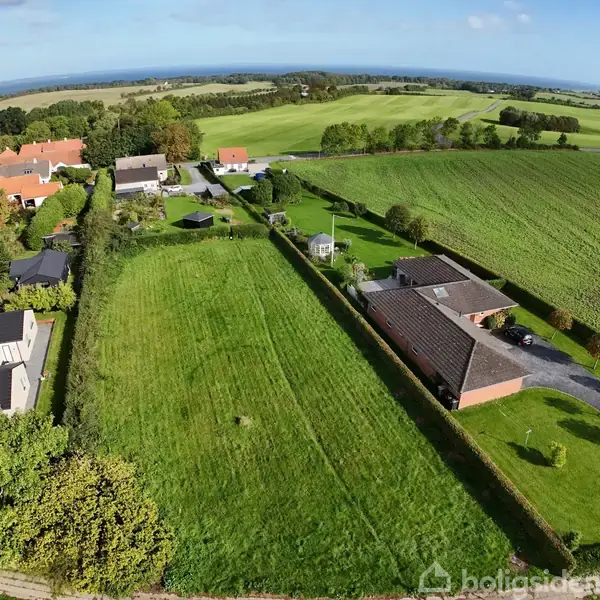 Hus ligger oven på en stor grøn mark, omgivet af lave hække og et mindre nabolag. Landbrugslandskabet strækker sig i baggrunden med veje og spredte træer under en blå himmel.