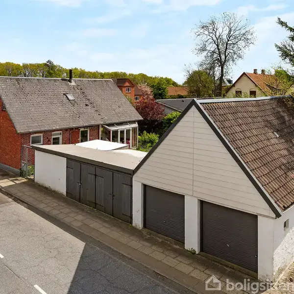Et rødt murstenshus står ved en vej, omgivet af andre huse og en garagebygning med tre porte. Træer og buske ses i baggrunden under en klar himmel.