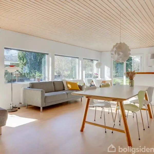 Stue med grå sofa og spisebord med stole, belyst af dagslys fra store vinduer. Et moderne køkken kan ses i baggrunden. Vegetation er synlig udenfor.