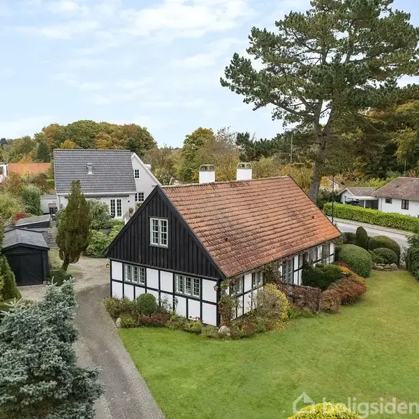 Et bindingsværkshus med rødt tegltag står i en frodig have omgivet af træer og buske. En grusvej fører op til huset, og nabohuse er synlige i baggrunden.