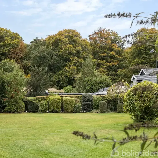 En grøn græsplæne omgivet af buske og træer i efterårsfarver, med et hus til højre. Flere små buske danner en hæk, og himlen er delvist overskyet.