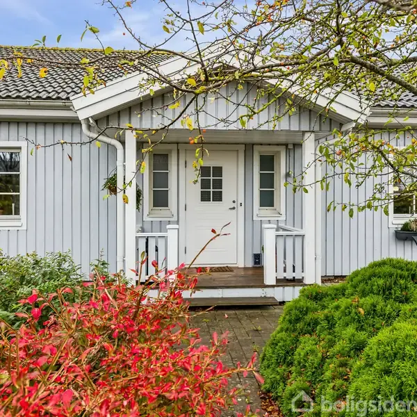 Hus står stille omkranset af farverige buske med røde blade og grøn beplantning. Husets facade er grå med hvidt dobbeltdør og små veranda.