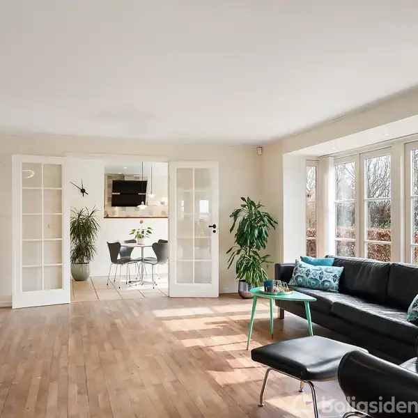 Stue med sort sofa og grønne puder står ved et stort vinduesparti, der giver udsigt til haven. En spisestue kan ses gennem to franske døre.