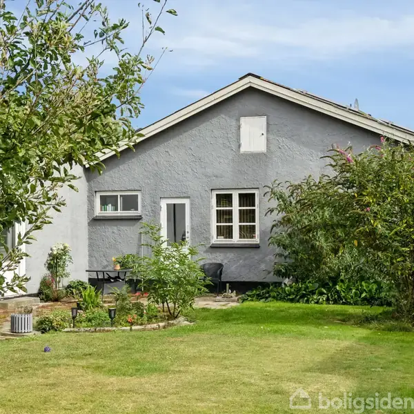 Et gråt hus med hvide vinduer står stille, omgivet af en grøn have med græs og buske under en blå himmel.