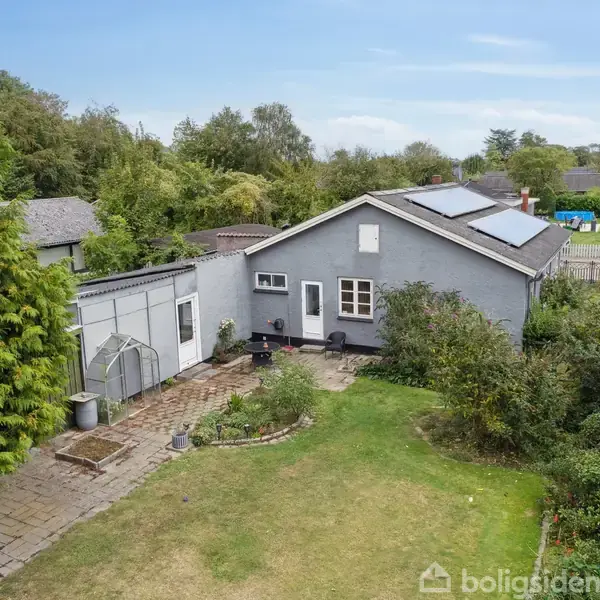 En grå étplansvilla med solpaneler på taget står i en frodig have med græsplæne og buske. Flisegang fører til huset, omgivet af træer og naboområder i baggrunden.