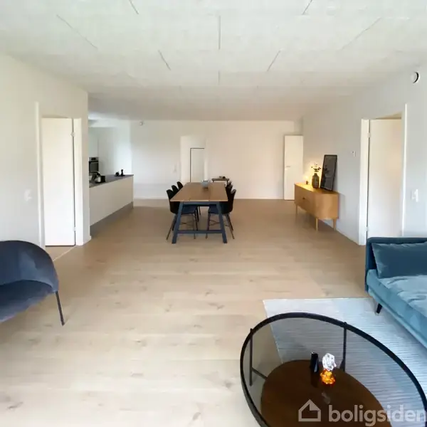 Et spisebord med sorte stole står i midten af en lys stue med trægulv. Til højre ses en blågrå sofa, og en skænk med dekorationer står langs væggen.