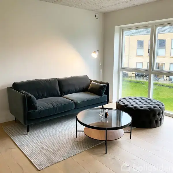 En sort sofa står i hjørnet af en lys stue ved siden af et rundt sofabord og en puf. En gulvlampe og store vinduer viser udsigt over en græsplæne.