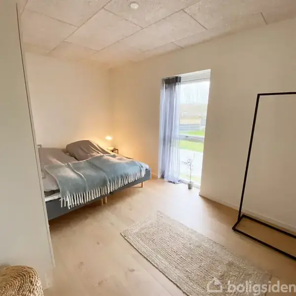 Sengen er redt, placeret ved siden af en natlampe på et lille bord, mod en lys væg. Et stort vindue med gardiner giver udsigt til en have.