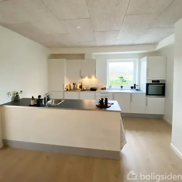 Et moderne køkken med hvidt inventar og sort bordplade. Køkkenet er udstyret med indbygget ovn, vask og vindue, som giver naturligt lys. Kryddere og planter dekorerer rummet.