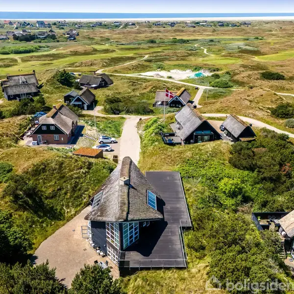 Stråtækte huse står spredt på et frodigt, bakket landskab. En sti snor sig gennem området, mens havet anes i horisonten. Området er præget af grønne marker og klitter.