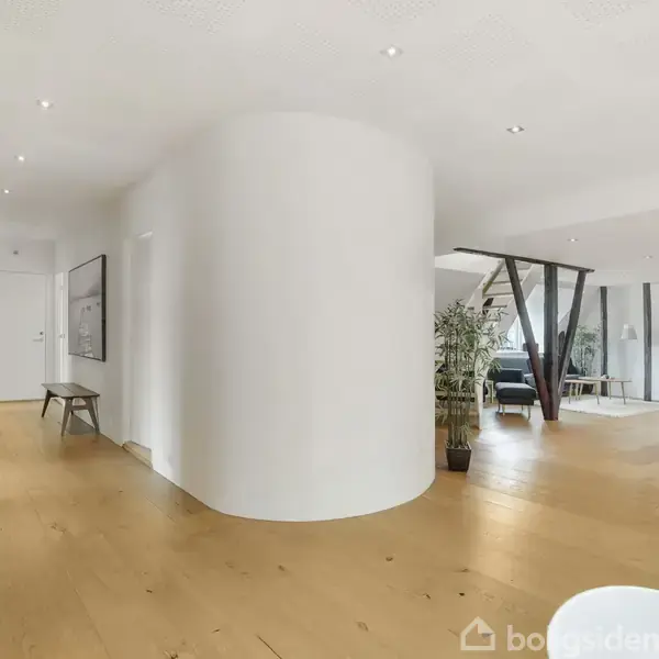 En stor, lys stue med træparketgulv og hvidmalede vægge. Rund, hvid søjle står centralt. Komfortable møbler og grønne planter ses i baggrunden. Ingen tekst til stede.