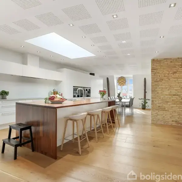 Køkken med stor køkkenø og barstole står i et lyst rum. En spiseplads er synlig i baggrunden, omgivet af lyse træ- og murstensvægge. Skarpt, moderne design.