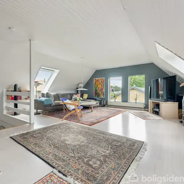 Et lyst loftstueområde med tæpper på gulvet viser en stor reol fyldt med bøger, en sofa, stole og et sofabord. Store vinduer og skråtag skaber en rummelig følelse.