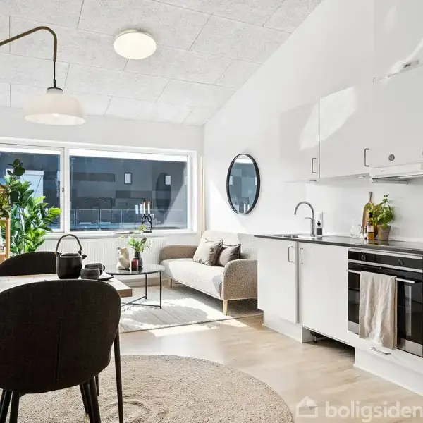 En stue og køkken i åben plan. Et spisebord har to stole foran og planter på bordet. En sofa og rundt spejl er modsat et stort vindue.