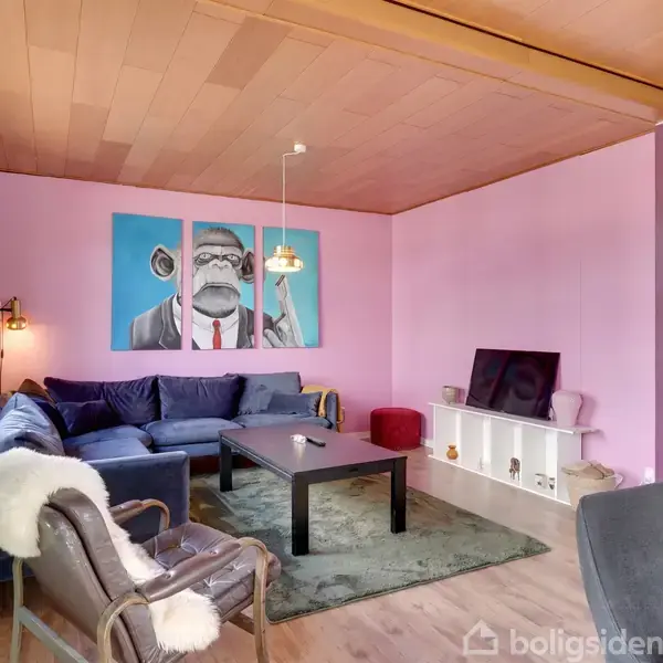 En stue med lilla vægge, blå sofa, og en lænestol med hvidt tæppe. Et stort maleri af en abe i jakkesæt hænger på væggen. En TV-enhed står ved siden af.