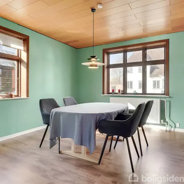 Spisebord med blå dug i midten af et rum, omringet af fire sorte stole. Grønne vægge og to vinduer tilbyder naturligt lys. En enkelt pendellampe hænger over bordet.