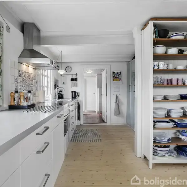 Køkken med hvide skabe, der er fuldt udstyret, herunder en emhætte og en åbne hylde fyldt med porcelæn. Vinduer til venstre slipper naturligt lys ind i det lyse rum.