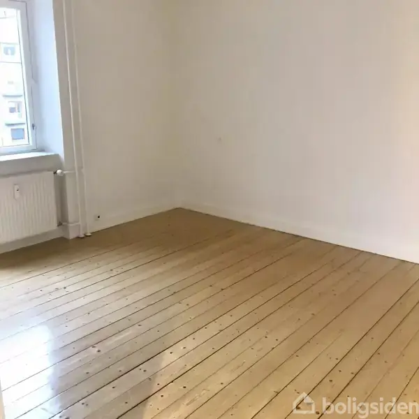 Et tomt værelse med lyse trægulve og hvide vægge. Mod venstre ses et vindue og en radiator, der indikerer en ydre væg. Ingen tekst.
