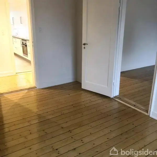 Et lyst rum med trægulv og to åbne døre. Den ene dør fører til et køkken med hvide skabe og en ovn. Ingen tekst fundet.