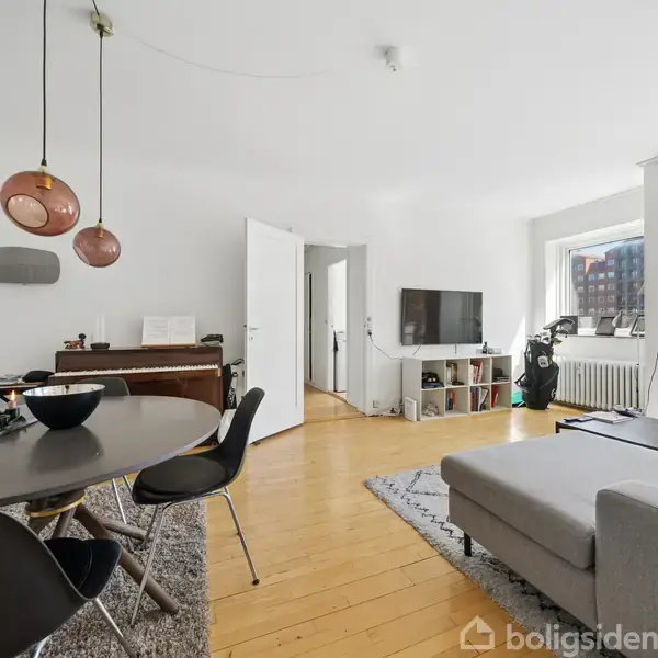 Lyst stue- og spisestueområde med rund spisebord, fire stole og en grå sofa. TV'et er placeret på en hylde. Store vinduer tillader naturligt lys at strømme ind.
