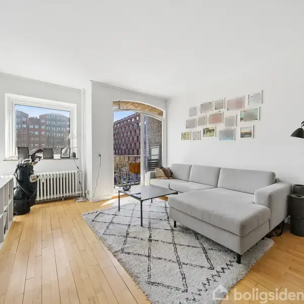 Lys stue med grå sofa, placeret ved et sofabord på et mønstret tæppe. Store vinduer og en altan giver udsigt til byens bygninger. En række små billeder hænger på væggen.