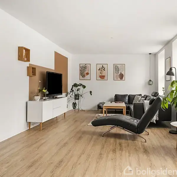 En moderne stue med en lænestol vendt mod et TV på et hvidt skab. Rummet har trægulv, grønne planter, en grå sofa og vægkunst. Store vinduer til venstre.