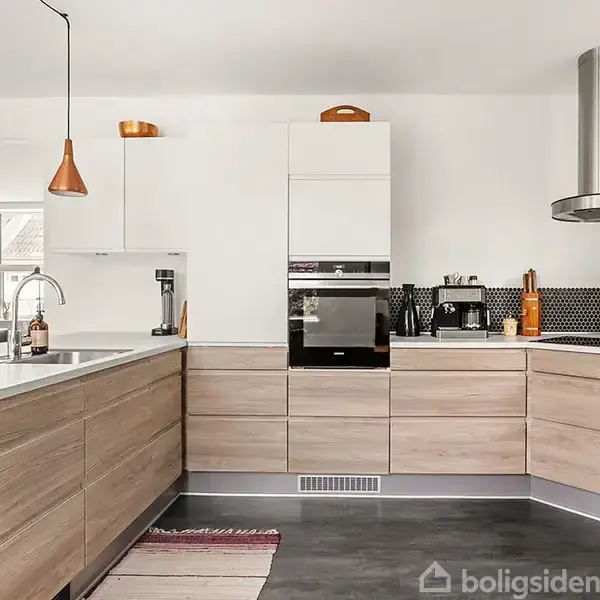 Køkken med træskabe og hvide bordplader, udstyret med ovn og emhætte. Kobberlamper hænger over vasken. Rummet har mørkt gulv og et lyst, moderne design.
