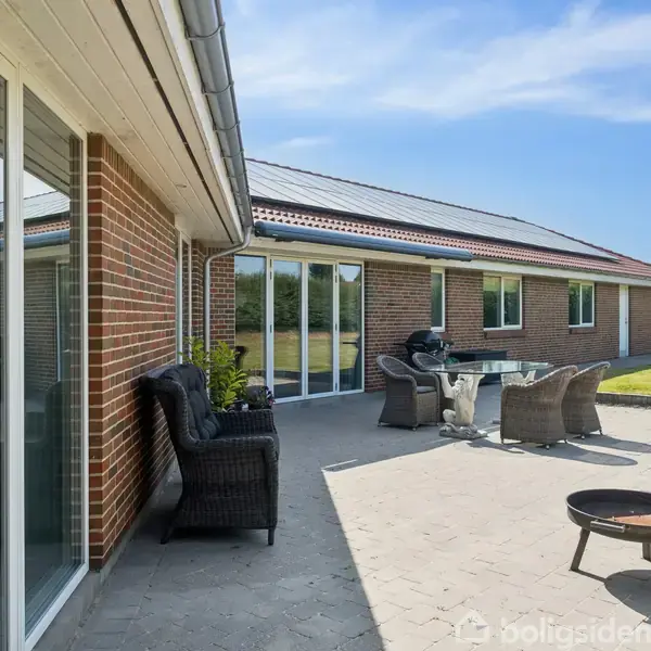 Murstenshus med terrassemøbler, herunder stole og bord, placeret på en flisebelagt terrasse. Omkranset af en velplejet græsplæne og under en klar, blå himmel.